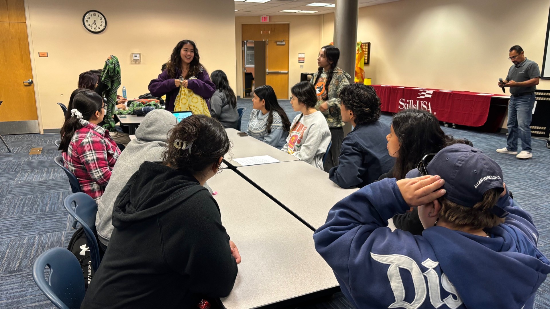 A group of student sitting at a table. One student is standing up and talking while holding a blanket. 